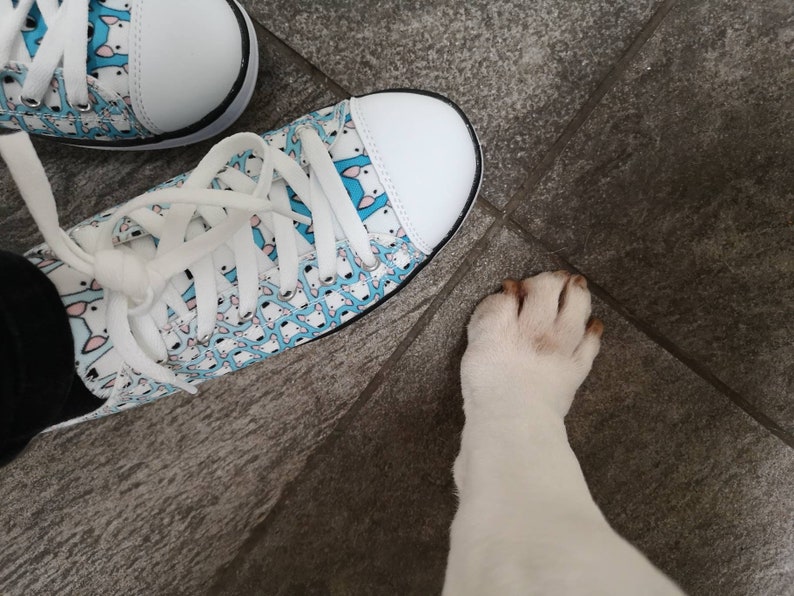 May include: A pair of white canvas shoes with a blue and white pattern of small dogs. The shoes are tied with white laces. A white dog's paw is visible in the lower right corner of the image.