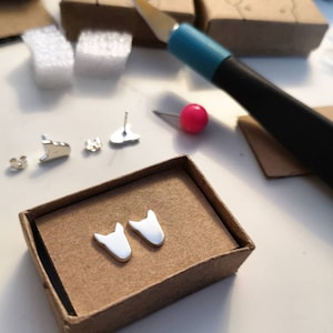 Puede incluir: Un par de pendientes de plata en forma de cabeza de perro, presentados en una pequeña caja marrón.