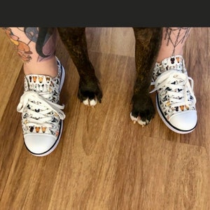 May include: A pair of white canvas shoes with a pattern of dog faces. The shoes are worn by a person with tattoos on their legs. The person's dog is standing on its hind legs with its paws on the person's shoes.