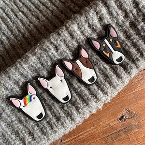 Może przedstawiać: Cztery kolorowe zawieszki z psem Bull Terrier o różnych wzorach i kolorach, w tym białym, brązowym i czarnym, ułożone na szarej dzianinowej powierzchni. Jedna zawieszka ma tęczowy wzór. Zawieszki umieszczone są na drewnianej powierzchni.