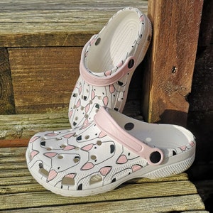May include: A pair of white clogs with a pink strap and a black and white print of bulldogs. The clogs are on a wooden surface.
