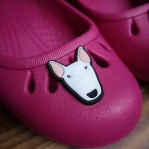 Puede incluir: Un par de bailarinas rosas con un dije de cara de perro blanco. El perro tiene ojos negros y una nariz negra.