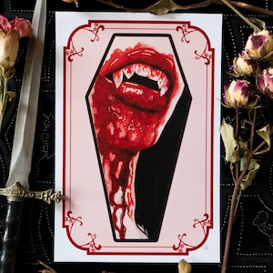 May include: A print featuring a vampire's mouth with sharp fangs, dripping with red blood, inside a coffin-shaped frame. The artwork has a gothic theme, with a red and black color scheme. Dried roses and a decorative dagger are in the background.
