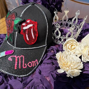 May include: A black baseball cap with rhinestones, a red sequined tongue patch, a pink rose patch, and the word "Mom" in pink rhinestones. The cap is on a purple background with a silver crown and white flowers.