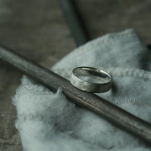 May include: A silver ring with a textured finish sits on a gray fabric surface. The ring is resting on a black metal rod.