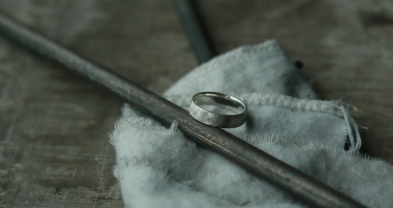 May include: A silver ring with a textured finish sits on a gray fabric surface. The ring is resting on a black metal rod.
