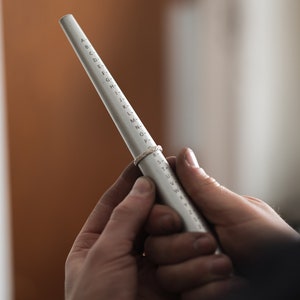 May include: A silver ring sizer tool with letters and numbers printed on it. A ring is placed on the sizer to determine the correct size. The tool is held by two hands, with a blurred background.