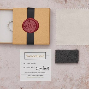 May include: A brown box with a black ribbon and a red wax seal with a white letter W. The box contains a silver ring. A white polishing cloth and a gray cleaning pad are also included. The box has a card with the text "WoodenGold" and the words "CRAFTED ON:" and "CRAFTSMAN: S.cichouls".