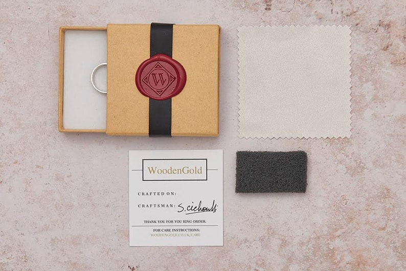 May include: A brown box with a black ribbon and a red wax seal with a white letter W. The box contains a silver ring. A white polishing cloth and a gray cleaning pad are also included. The box has a card with the text "WoodenGold" and the words "CRAFTED ON:" and "CRAFTSMAN: S.cichouls".