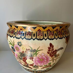 May include: A large, round, white ceramic planter with a gold, black and red floral design. The planter has a wide rim and a slightly flared lip.