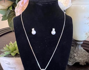 May include: A silver necklace and matching earrings set with a pearl pendant and pearl studs. The jewelry is displayed on a black velvet bust, with a floral arrangement in the background. The necklace chain is delicate.