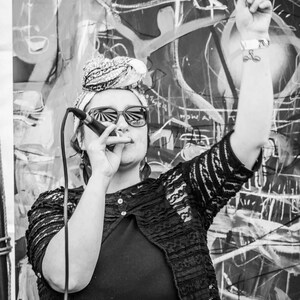 May include: A woman wearing a black lace top and a headscarf sings into a microphone. She is wearing sunglasses and has her arm raised in the air.