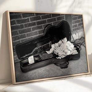 Può includere: Stampa in bianco e nero di una custodia per chitarra aperta traboccante di banconote in dollari USA. La custodia è appoggiata su una superficie di cemento contro un muro di mattoni.