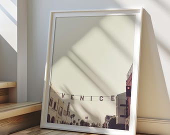 May include: A framed photograph of a street scene in Venice, California. The word "VENICE" is spelled out in red letters across the top of the image. The buildings in the background are a pale pink color.