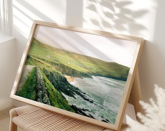 May include: A framed landscape print featuring a coastal scene with a road, stone wall, and ocean waves. The print has a light wood frame and is propped on a woven chair. The image showcases green hills and a sandy beach.