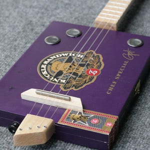 May include: A purple cigar box guitar featuring a Knuckle Sandwich logo with a skull graphic, three metal sound holes, a wooden bridge with a threaded metal rod, and a light wood neck. The text Chef Special is printed on the purple box surface.