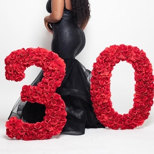 May include: A woman in a black dress stands beside large red floral numbers spelling out "30". The dress features a textured bodice and a flowing tulle skirt. The numbers are made of red roses, creating a celebratory theme.