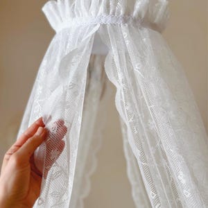 以下が含まれることがあります： 繊細な花柄とフリル付きのトップエッジが特徴の白いレースのキャノピー。透け感のある生地が中心から吊り下げられており、複雑な半透明のメッシュ素材のパネルが重なり合ってドレープ効果を生み出しています。
