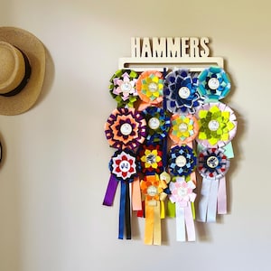 May include: A wooden wall display rack with the name HAMMERS cut out at the top, holding a collection of colorful fabric award rosettes. The rosettes feature various pleated designs in shades of green, orange, blue, yellow, purple, and pink, each with long hanging ribbons attached to the bottom.