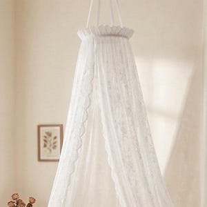 May include: A white, sheer, floor-length bed canopy made of delicate lace fabric. The canopy features a circular top with a ruffled edge, suspended from the ceiling, and drapes down with scalloped edges along the opening, creating a soft, layered effect.