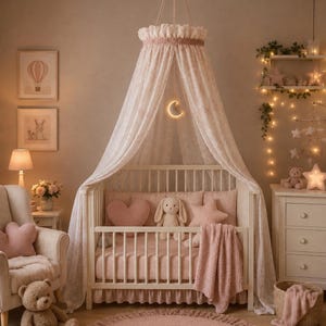 May include: A white wooden crib featuring a sheer, ruffled canopy draped from the ceiling. The crib is styled with soft pink bedding, a heart-shaped pillow, a star-shaped pillow, and a plush rabbit toy, illuminated by a glowing crescent moon light.