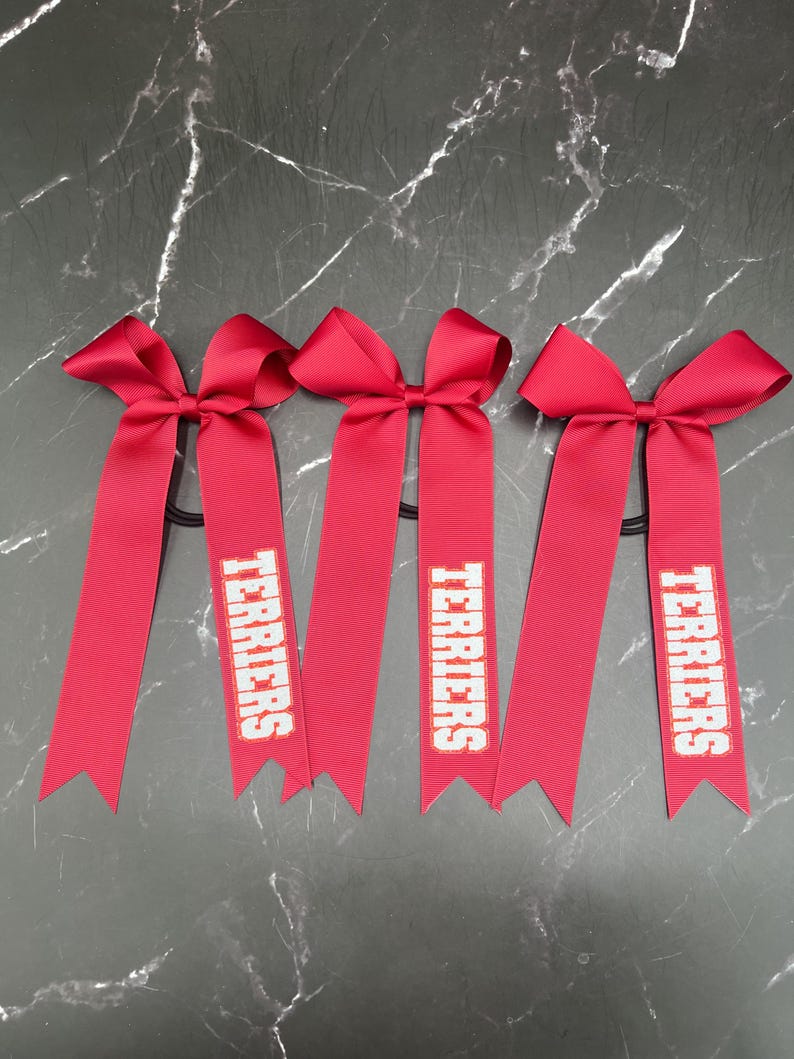 May include: Four red hair bows with long, ribbon tails. Each tail has the word "TERRIERS" printed in white. The bows are tied to black elastic hair ties, perfect for cheerleading or school spirit.