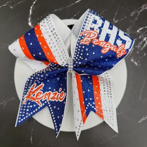 Op de afbeelding: Een cheerleading strik met een witte basis, blauwe en oranje strepen en sprankelende strass-steentjes. De strik is versierd met de tekst "BHS Bengals" en de naam "Kenzie" in oranje script. De strik is ontworpen voor cheerleading.