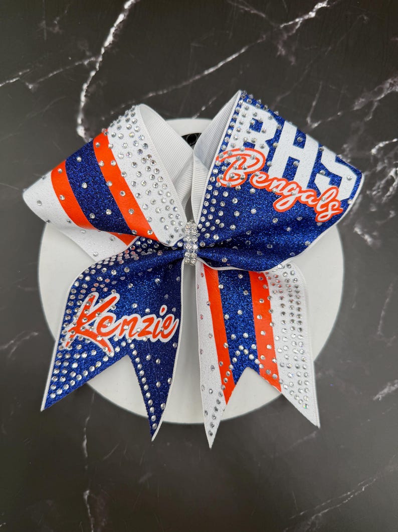 Op de afbeelding: Een cheerleading strik met een witte basis, blauwe en oranje strepen en sprankelende strass-steentjes. De strik is versierd met de tekst "BHS Bengals" en de naam "Kenzie" in oranje script. De strik is ontworpen voor cheerleading.