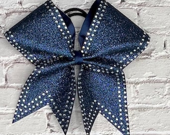 May include: A navy blue glitter cheerleading bow with a row of small, clear rhinestones outlining the edges. The bow is attached to a black elastic hair tie. The bow is a classic accessory for cheerleaders and dancers.