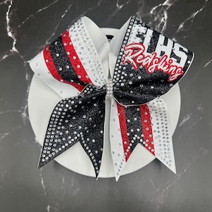 Op de afbeelding: Een cheerleading strik met een witte basis, zwarte en rode accenten en sprankelende strass-steentjes. De strik heeft de tekst "EHHS Redskins" in rood en wit. De strik is gecentreerd met een strass sluiting.