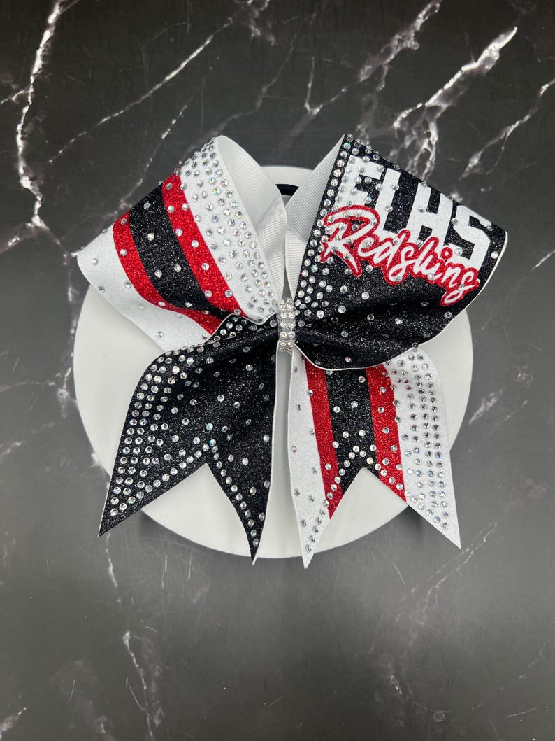 Op de afbeelding: Een cheerleading strik met een witte basis, zwarte en rode accenten en sprankelende strass-steentjes. De strik heeft de tekst "EHHS Redskins" in rood en wit. De strik is gecentreerd met een strass sluiting.