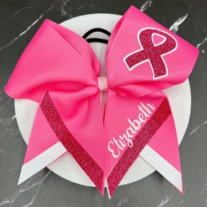 May include: Bright pink ribbon hair bow with a glitter ribbon accent and the name "Elizabeth" in white script. The bow features a pink ribbon with a white outline of a ribbon symbol.