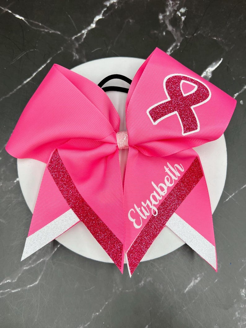 May include: Bright pink ribbon hair bow with a glitter ribbon accent and the name "Elizabeth" in white script. The bow features a pink ribbon with a white outline of a ribbon symbol.