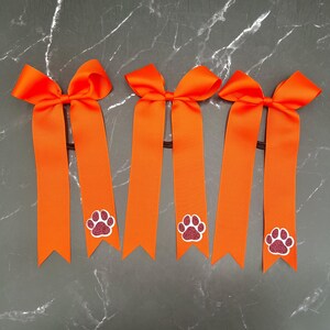 May include: Five orange hair bows with long, ribbon tails. Each bow features a glittery paw print design in the center of the ribbon. The bows are attached to black elastic hair ties.