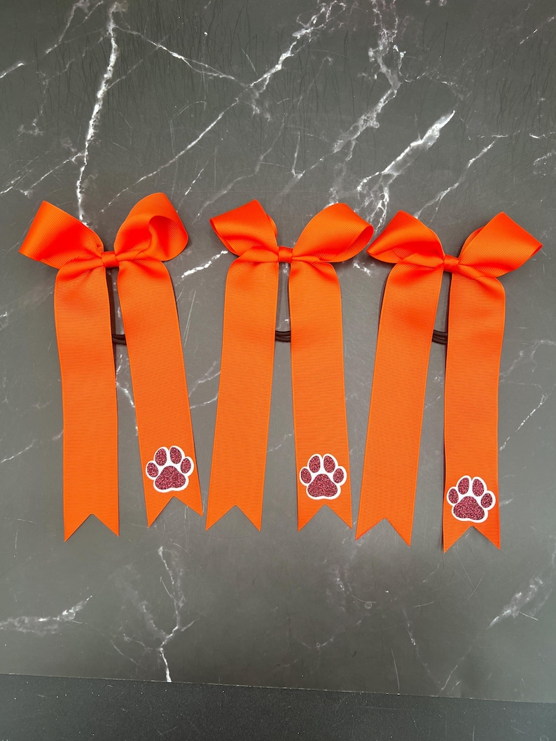 May include: Five orange hair bows with long, ribbon tails. Each bow features a glittery paw print design in the center of the ribbon. The bows are attached to black elastic hair ties.