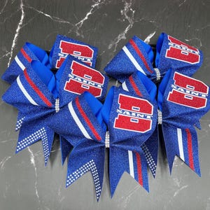 May include: A collection of blue glitter cheerleading bows with red and white accents. Each bow features the word "SAINT" in red block letters. The bows are adorned with rhinestones and have a pointed tail design.