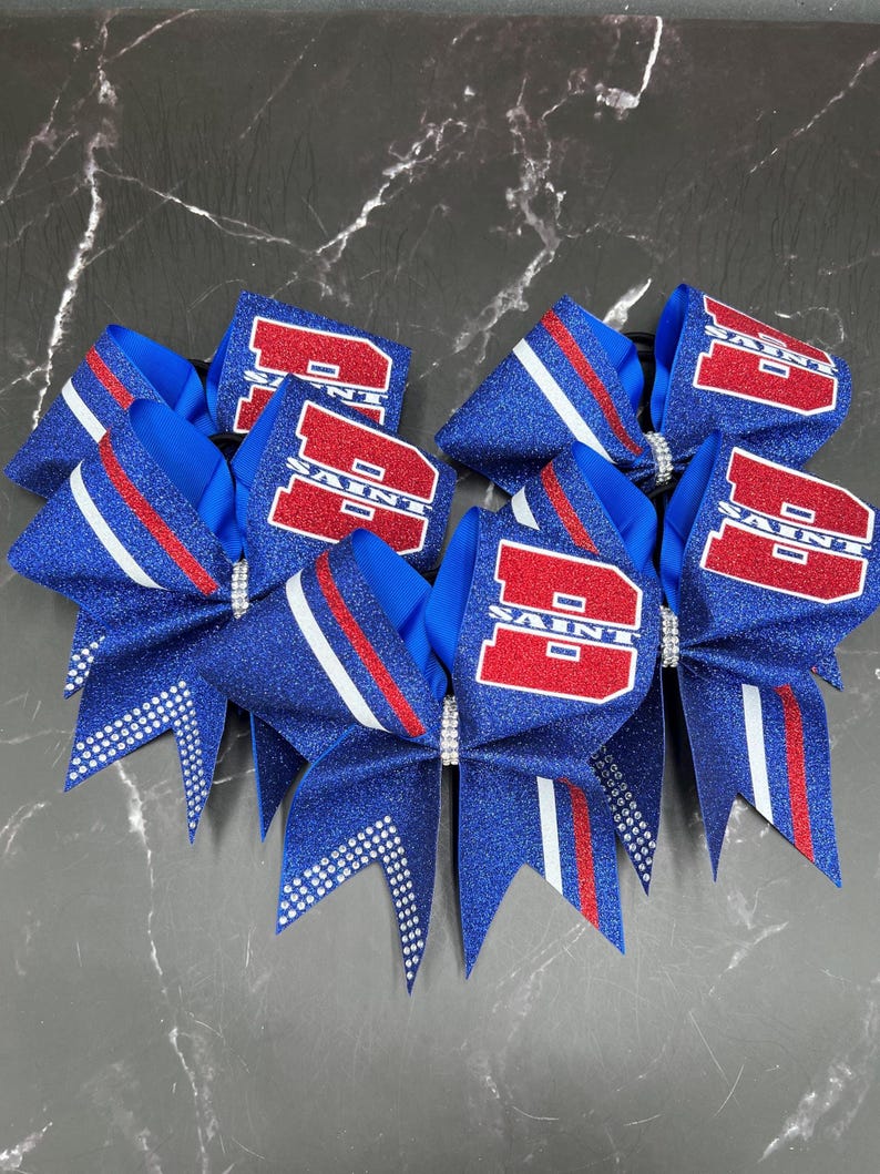 May include: A collection of blue glitter cheerleading bows with red and white accents. Each bow features the word "SAINT" in red block letters. The bows are adorned with rhinestones and have a pointed tail design.