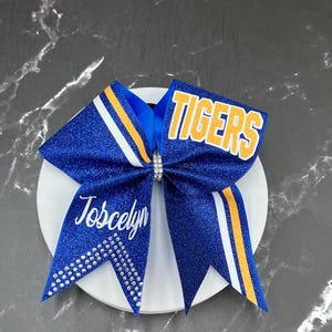 May include: A blue glitter cheerleading bow with the word "TIGERS" in yellow and white. The bow has white and yellow stripes and the name "Joscelyn" in white script. Rhinestones adorn the bow.