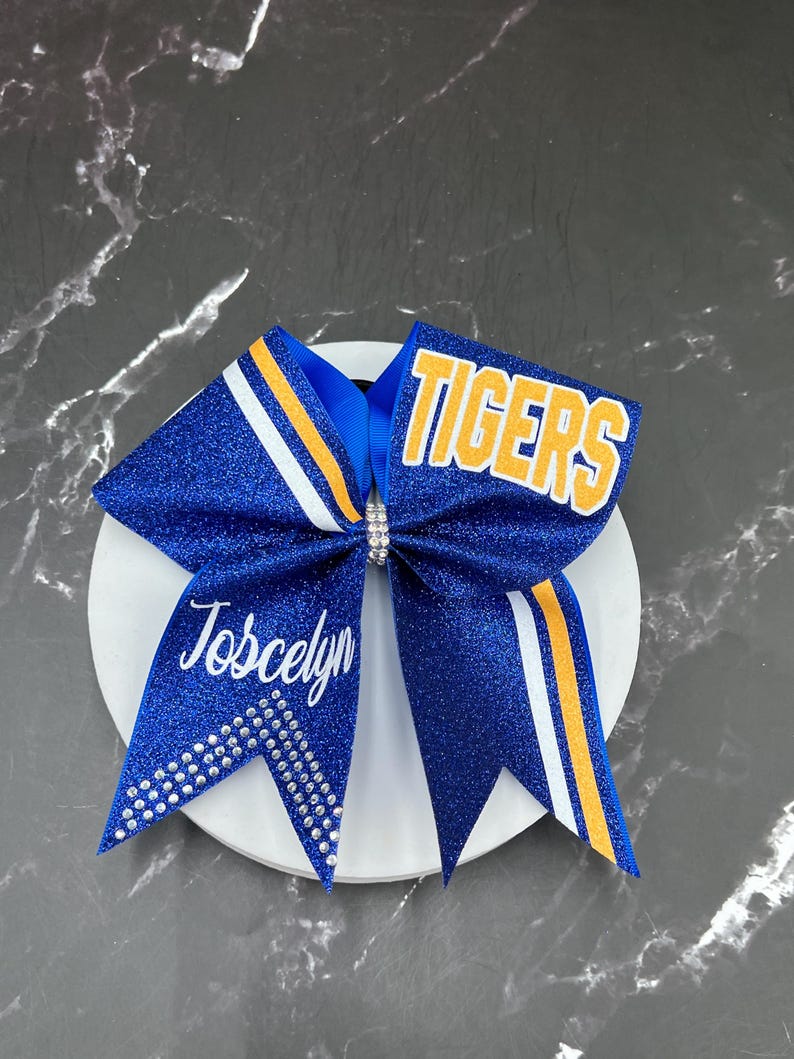 May include: A blue glitter cheerleading bow with the word "TIGERS" in yellow and white. The bow has white and yellow stripes and the name "Joscelyn" in white script. Rhinestones adorn the bow.