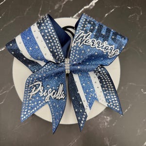 Op de afbeelding: Een blauw-witte cheerleading strik met het woord "Warriors" en de naam "Priscilla" in zilveren letters. De strik heeft glitter- en strassaccenten en is bevestigd aan een zwarte elastische band.