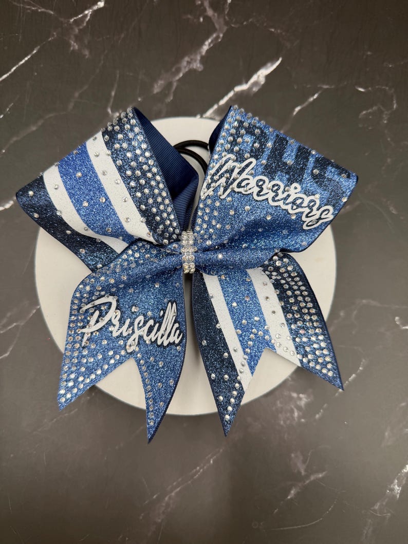 Op de afbeelding: Een blauw-witte cheerleading strik met het woord "Warriors" en de naam "Priscilla" in zilveren letters. De strik heeft glitter- en strassaccenten en is bevestigd aan een zwarte elastische band.