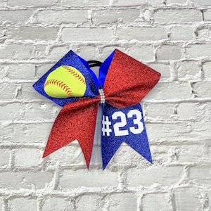 May include: A softball-themed hair bow with a yellow softball graphic on a blue background, red glitter accents, and the number 23 in white. The bow is embellished with a rhinestone center and is set against a white brick wall.