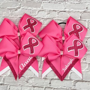 May include: Pink cheerleading bows with a glitter ribbon design and the text "Anali" and "Takayla". Each bow features a pink ribbon design with a glitter ribbon. The bows are on a white brick background.