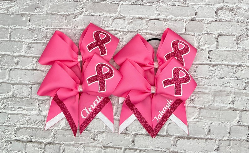 May include: Pink cheerleading bows with a glitter ribbon design and the text "Anali" and "Takayla". Each bow features a pink ribbon design with a glitter ribbon. The bows are on a white brick background.