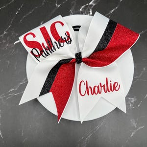May include: A cheer bow featuring white, red glitter, and black glitter ribbons. The left side displays the red text SJC and black script text Panthers. The right side features the red script name Charlie. A black glitter center knot holds the bow.