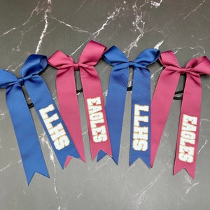 May include: Six satin hair bows in blue and maroon. Each bow has a long ribbon with white text. The text reads "LLHS" and "EAGLES". The bows are arranged on a dark gray marble surface.