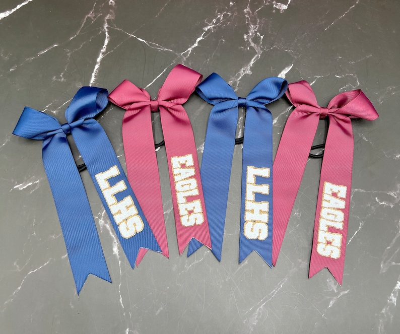 May include: Six satin hair bows in blue and maroon. Each bow has a long ribbon with white text. The text reads "LLHS" and "EAGLES". The bows are arranged on a dark gray marble surface.