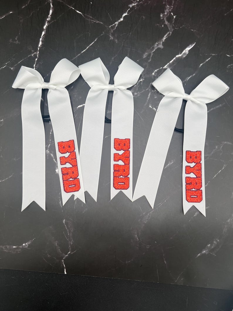 May include: Four white hair bows with red glitter text that reads "BYRD". Each bow has a ribbon tail and is attached to a black elastic band. The bows are arranged on a black and white marble surface.