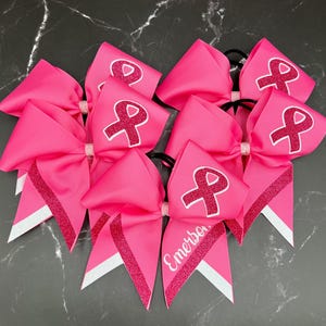 May include: Bright pink cheerleading bows with a white ribbon design and glitter accents. One bow has the name "Emerson" in white script. These bows are perfect for supporting a cause.