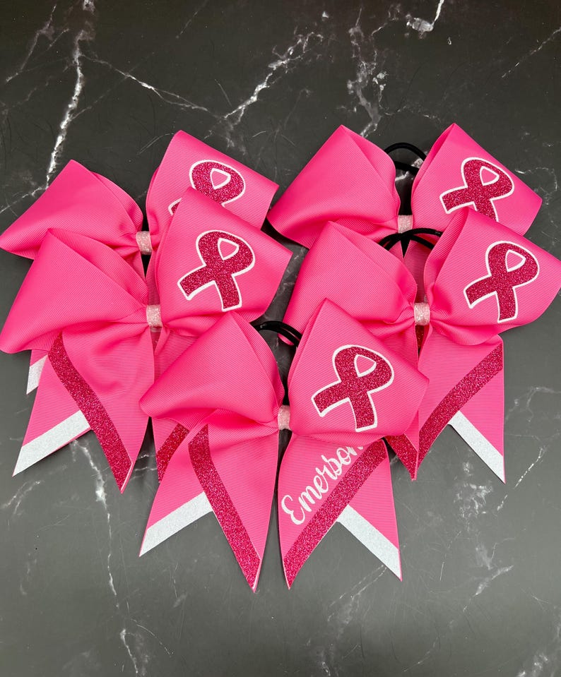 May include: Bright pink cheerleading bows with a white ribbon design and glitter accents. One bow has the name "Emerson" in white script. These bows are perfect for supporting a cause.